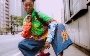 Jeune femme noire, accroupie dans une rue sur le trottoir, habillé avec un jean pâtes d'éléphant et un sweat très coloré.