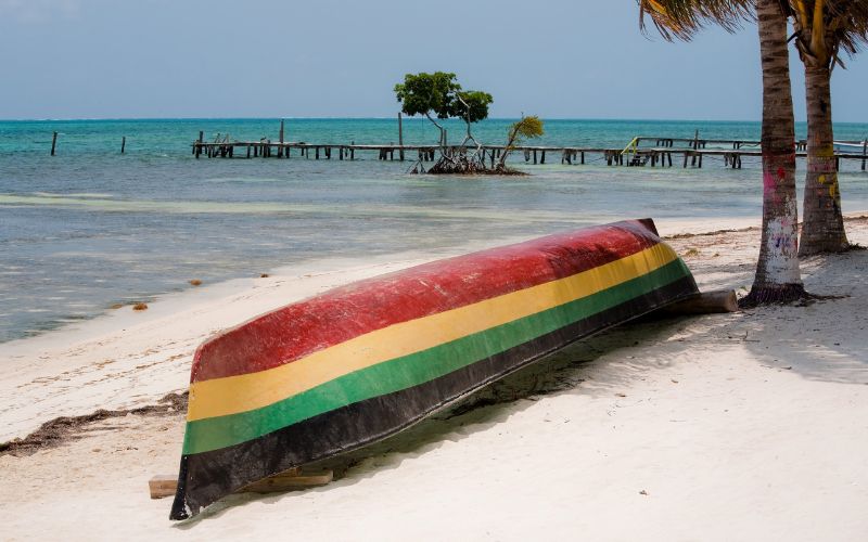 Barque en bois, retournée et peinte aux couleurs de la Jamaïque : rouge, jaune, vert, noir. Sur une plage de sable blanc, on voit la mer et des palmiers en fond.