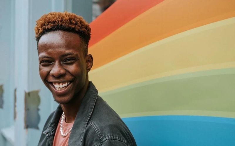Jeune homme avec un grand sourire, portant un t-shirt rouge dรฉlavรฉ et une veste en jean gris-noir, il pose devant un mur avec des bandes de couleurs peintes comme un arc-en-ciel.