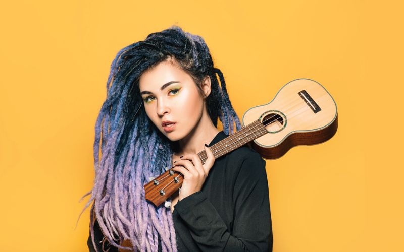 Femme avec des rastas logs colorรฉs noir, bleu et violet en dรฉgradรฉ. Elle porte un sweat noir et tien avec sa main un ukulรฉlรฉ posรฉ sur son รฉpaule. Un fond jaune derriรจre elle.