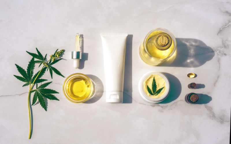Vu d'en haut : une tige de plante de CBD, de l'huile dans un pot, une crรจme, un baume, de l'huile dans un flacon verre, et des gรฉlules, chaque produit est ร base de CBD.