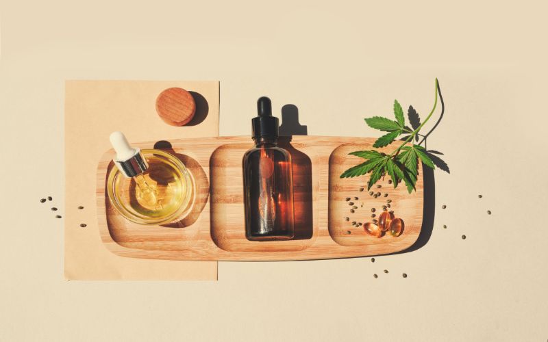 Plateau en bois rectangulaire, avec du CBD sous diffรฉrentes formes, de l'huile, des gรฉlules, des graines, des feuilles, vu d'en haut.