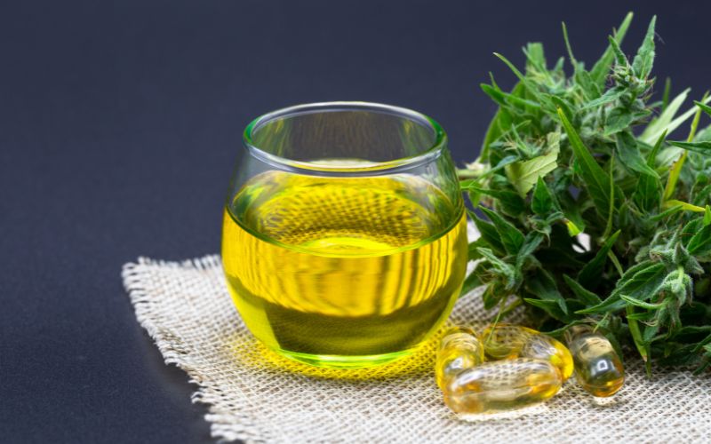 Verre d'infusion de chanvre posรฉ sur de la toile de jute, ร cรดtรฉ d'une fleur de canabis et de gรฉlule de CBD.
